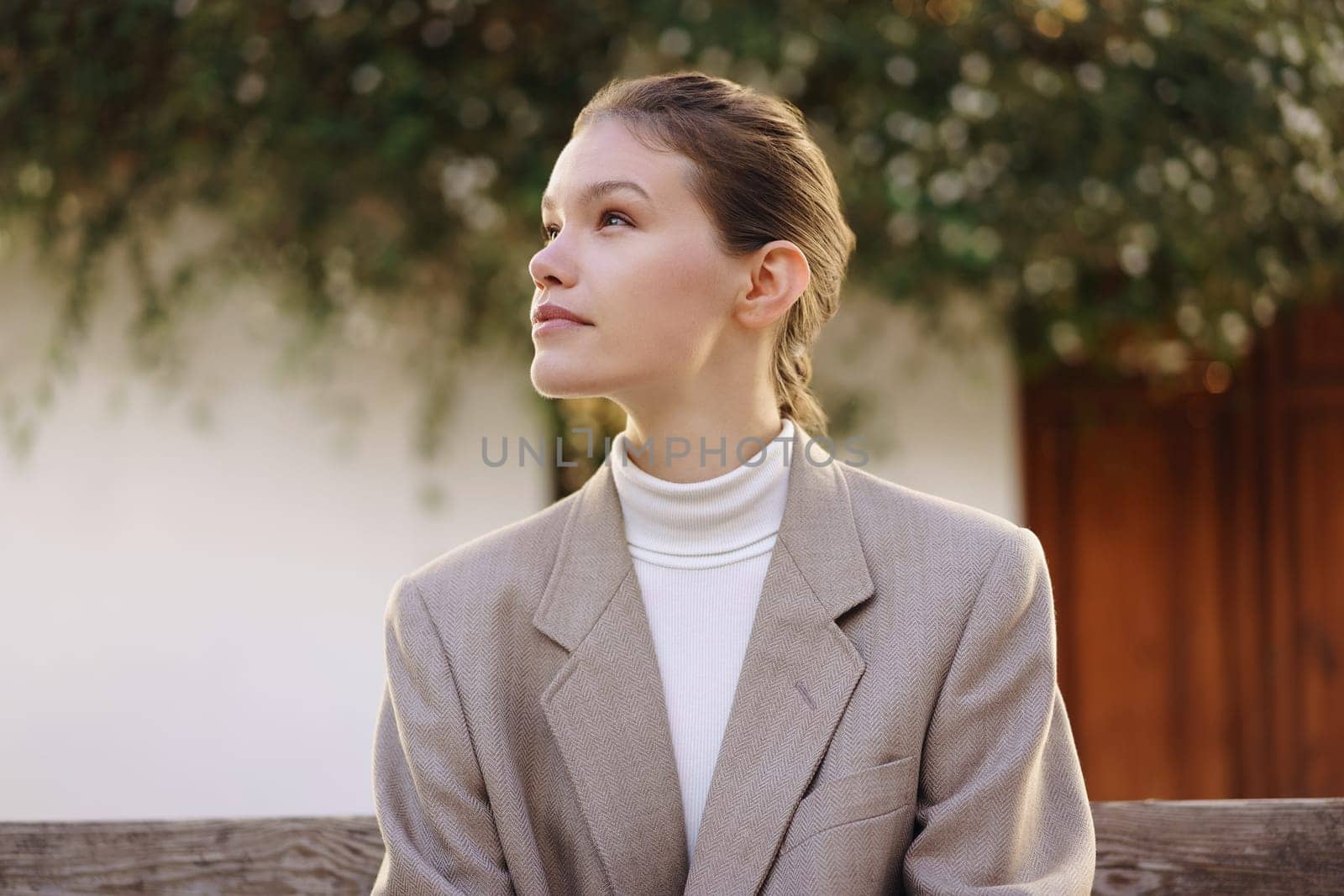 A young woman in a chic blazer and stylish turtleneck gazes thoughtfully outdoors, surrounded by lush greenery, radiating a calming and serene vibe as she enjoys the peaceful scenery