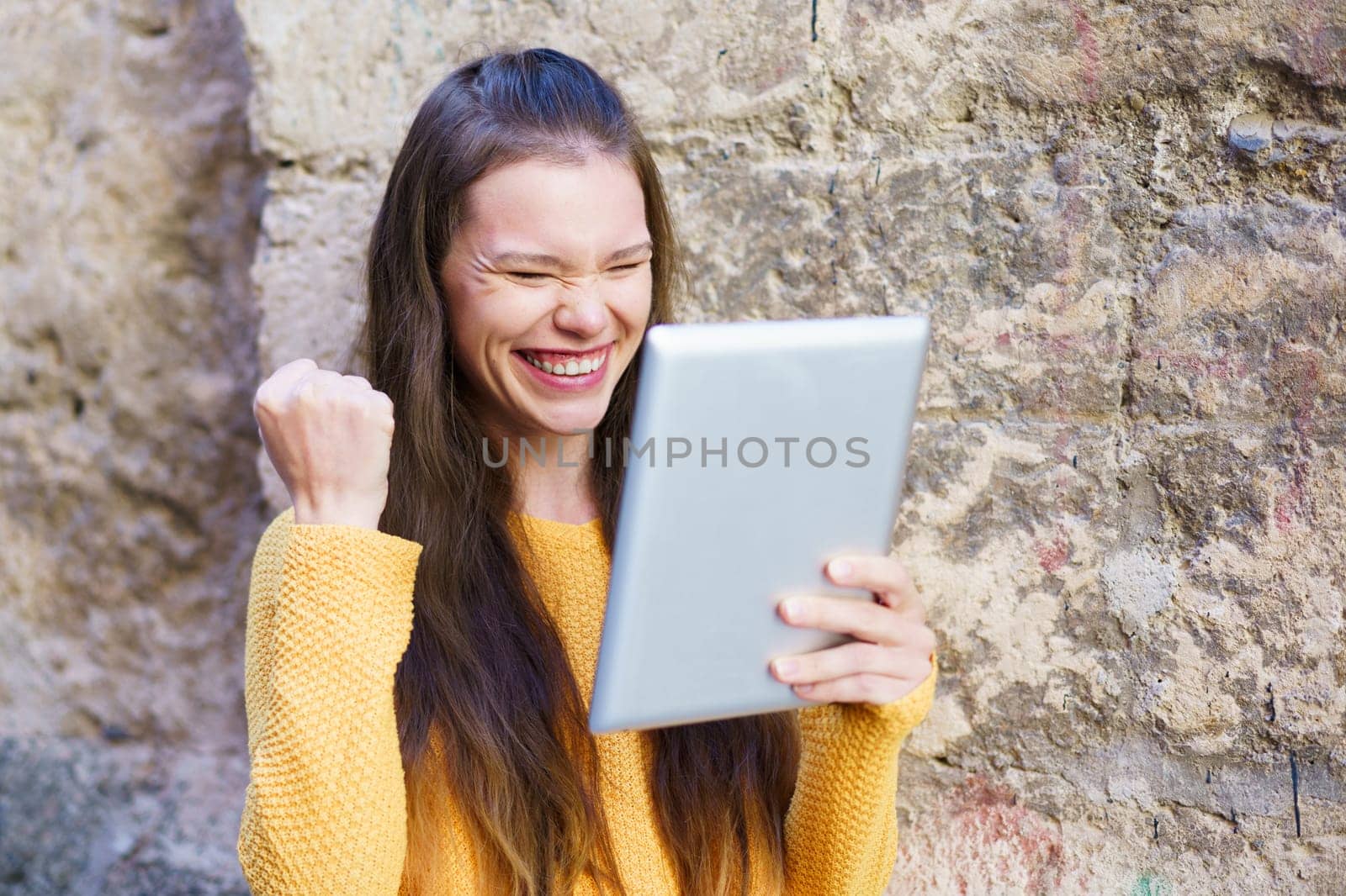 A young woman with long hair celebrates her achievements, smiling at her tablet. The bright, sunny day enhances her cheerful mood, creating a vibrant atmosphere full of positive energy