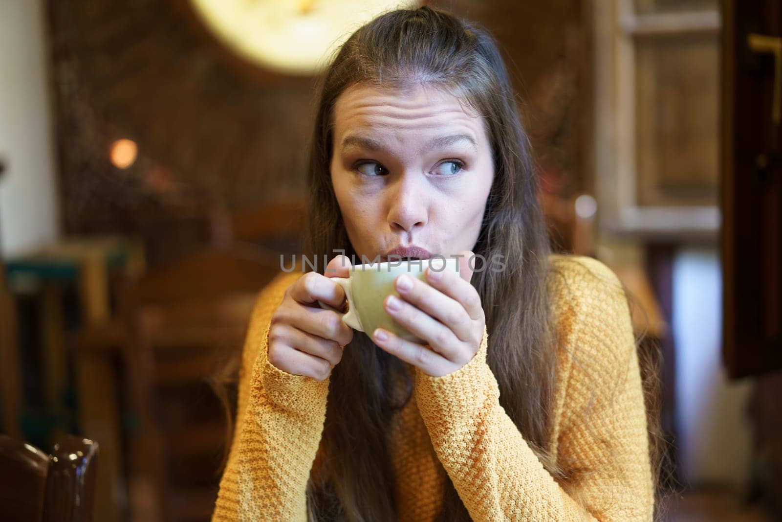 A young woman in a cozy caf sips coffee, lost in thought. She wears a warm sweater, embodying a moment of relaxation and introspection