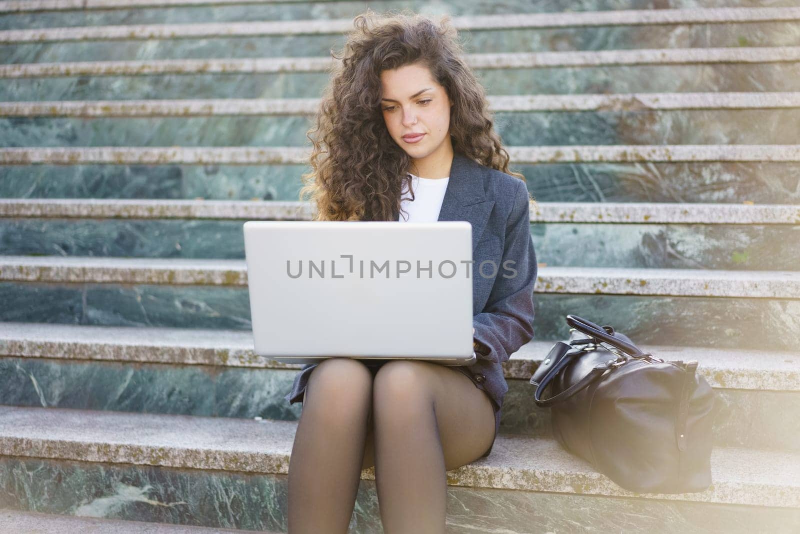 A young professional with curly hair is sitting on outdoor steps, focused on her laptop. She is dressed smartly, with a stylish bag beside her, embodying productivity and modern work culture.