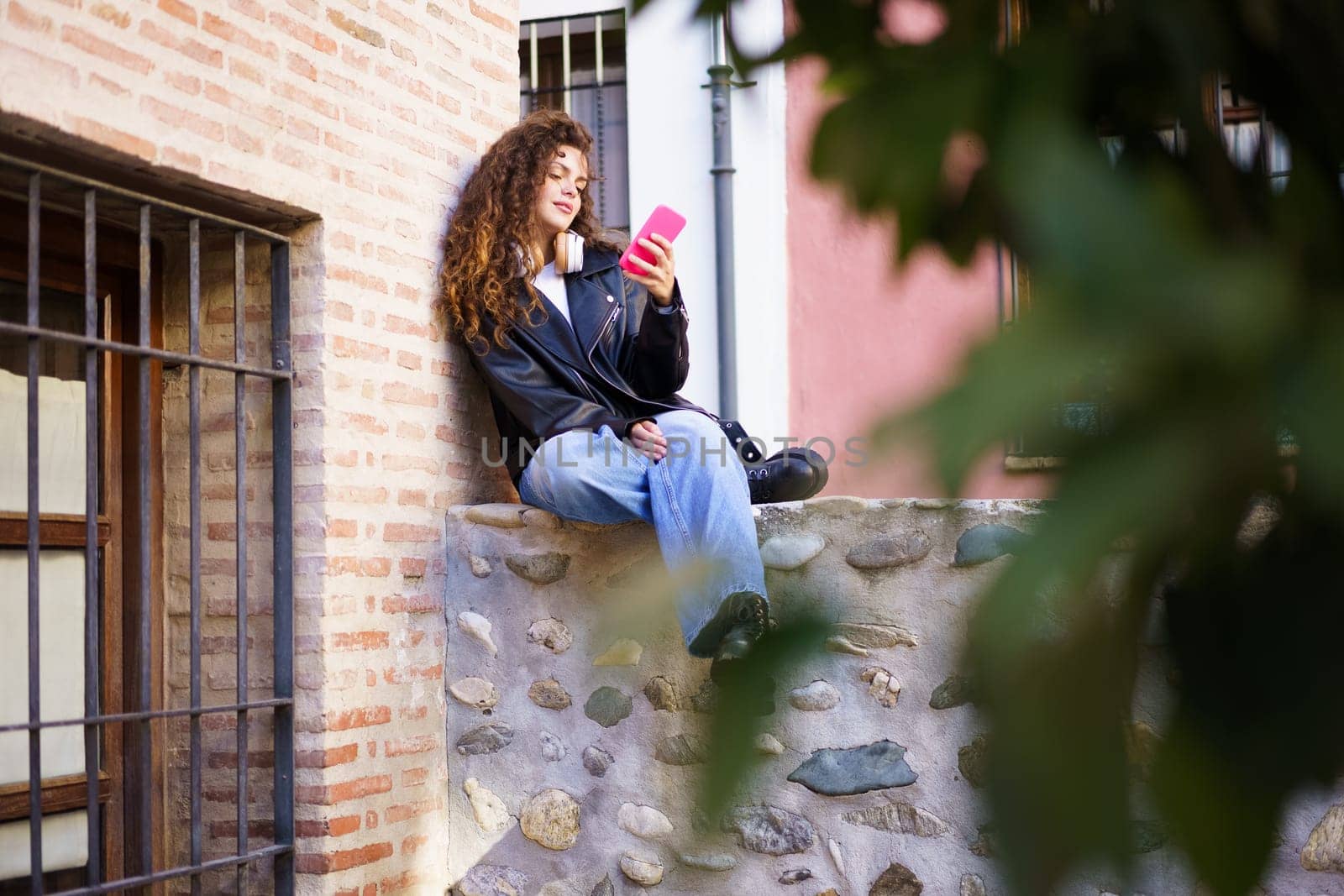 In a busy urban setting, a young woman with curly hair sits on a stone wall, focused on her smartphone, surrounded by vibrant green foliage, blending modern life with natures tranquility