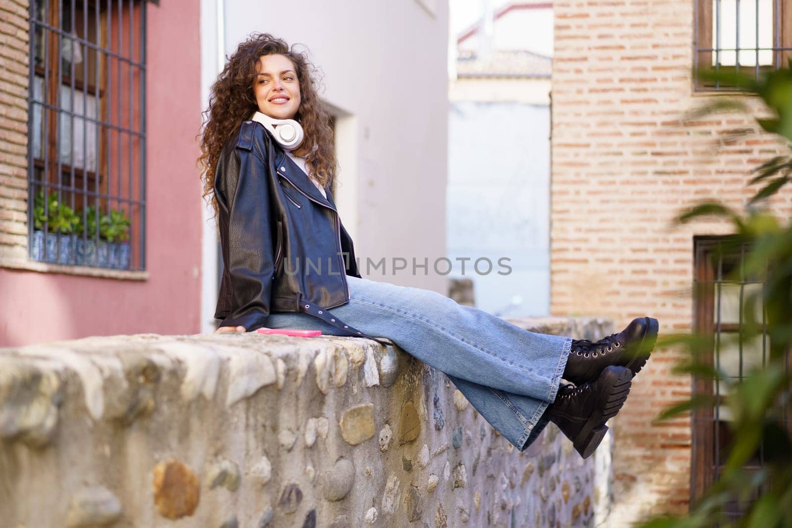 In a lively and charming urban backdrop, a fashionable young woman with naturally curly hair, dressed in a trendy leather jacket, happily enjoys her leisure time, sitting comfortably on a stone wall