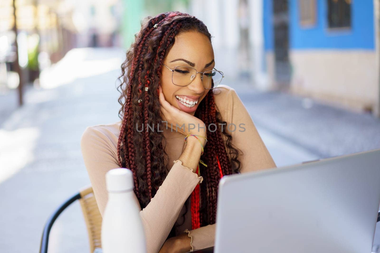A cheerful and joyful young woman with beautiful braided hair is working diligently on her laptop while sitting outside, fully embracing and enjoying the bright sunny atmosphere around her
