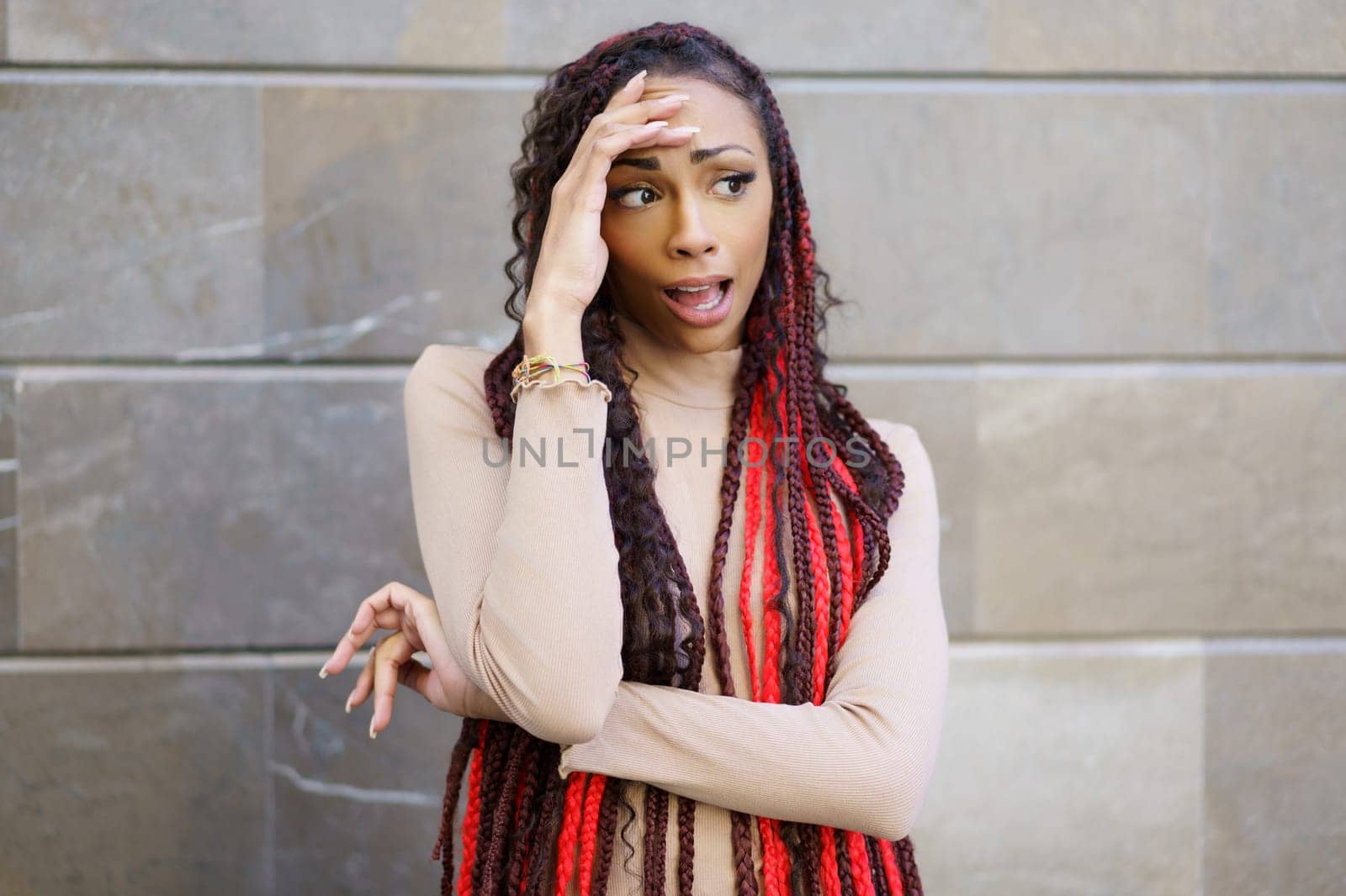 A stylish woman proudly displays her vibrant red and black dreadlocks while striking a thoughtful pose against a modern wall backdrop, which perfectly complements and frames her chic look