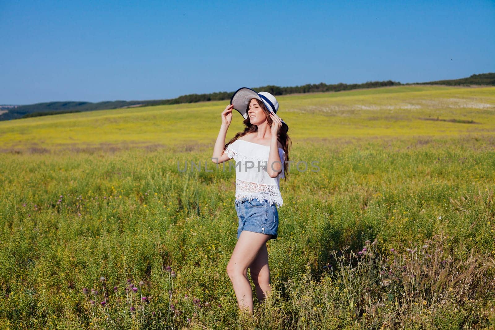 Woman in summer hat with brim smiling and walking in rural field in summer by road