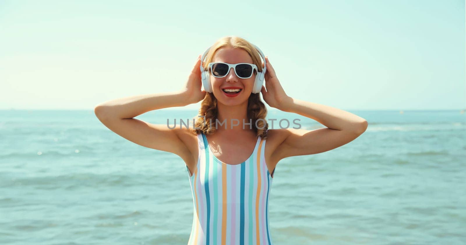 Summer vacation of happy young woman smiles enjoying listening to music with headphones, relaxed cheerful girl wearing a sunglasses on the beach at sea coast with blue sky on a sunny day