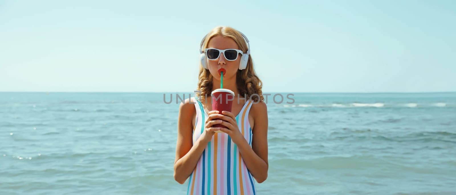 Summer vacation of happy relaxed young woman with cup of fresh juice drink enjoying listening to music with headphones, modern girl wearing a sunglasses on the beach at sea coast background