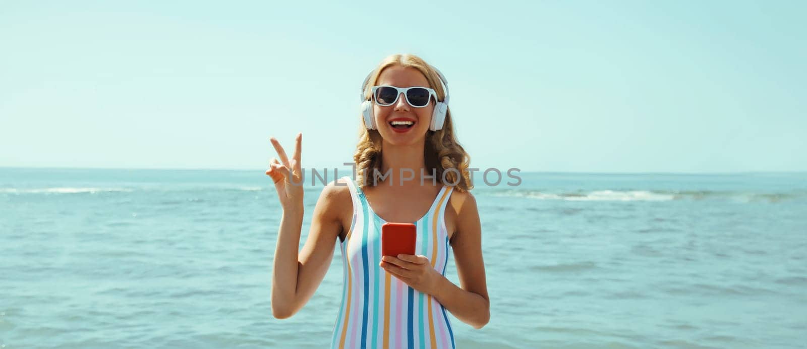 Summer vacation of happy young woman smiles enjoying listening to music with headphones, holding phone, relaxed cheerful girl wear a sunglasses on beach at sea coast with blue sky on a sunny day