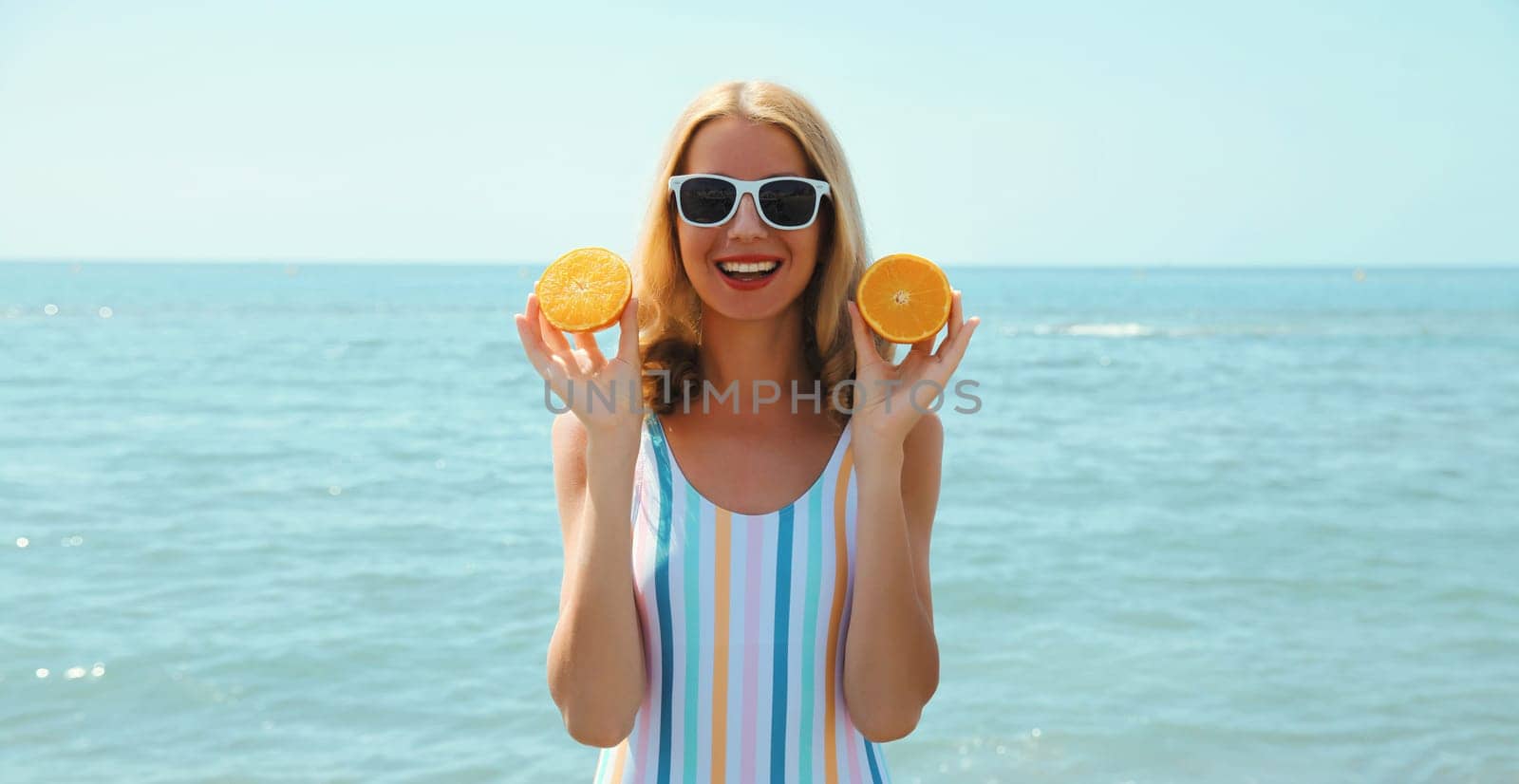 Summer vacation, portrait of happy smiling young woman, laughing, holding oranges fruits, having fun, wearing a swimsuit on the beach on sea coast background