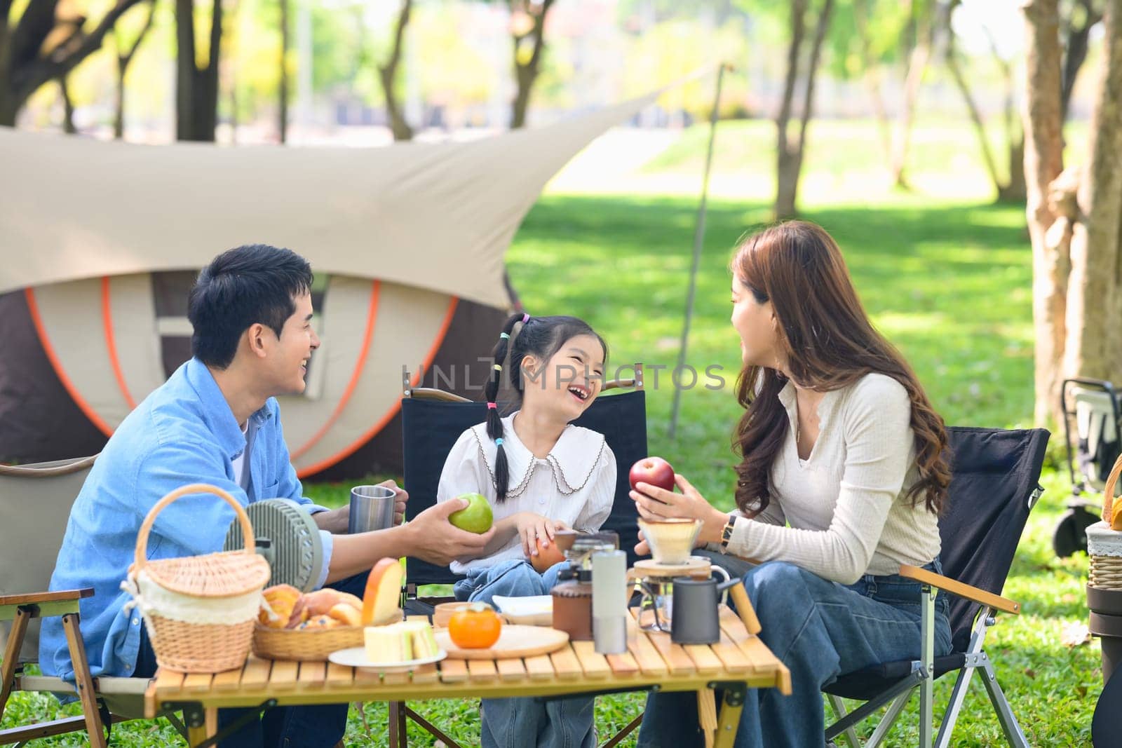 Parents and joyful daughter having a relaxing outdoor meal during family camping trip in nature.
