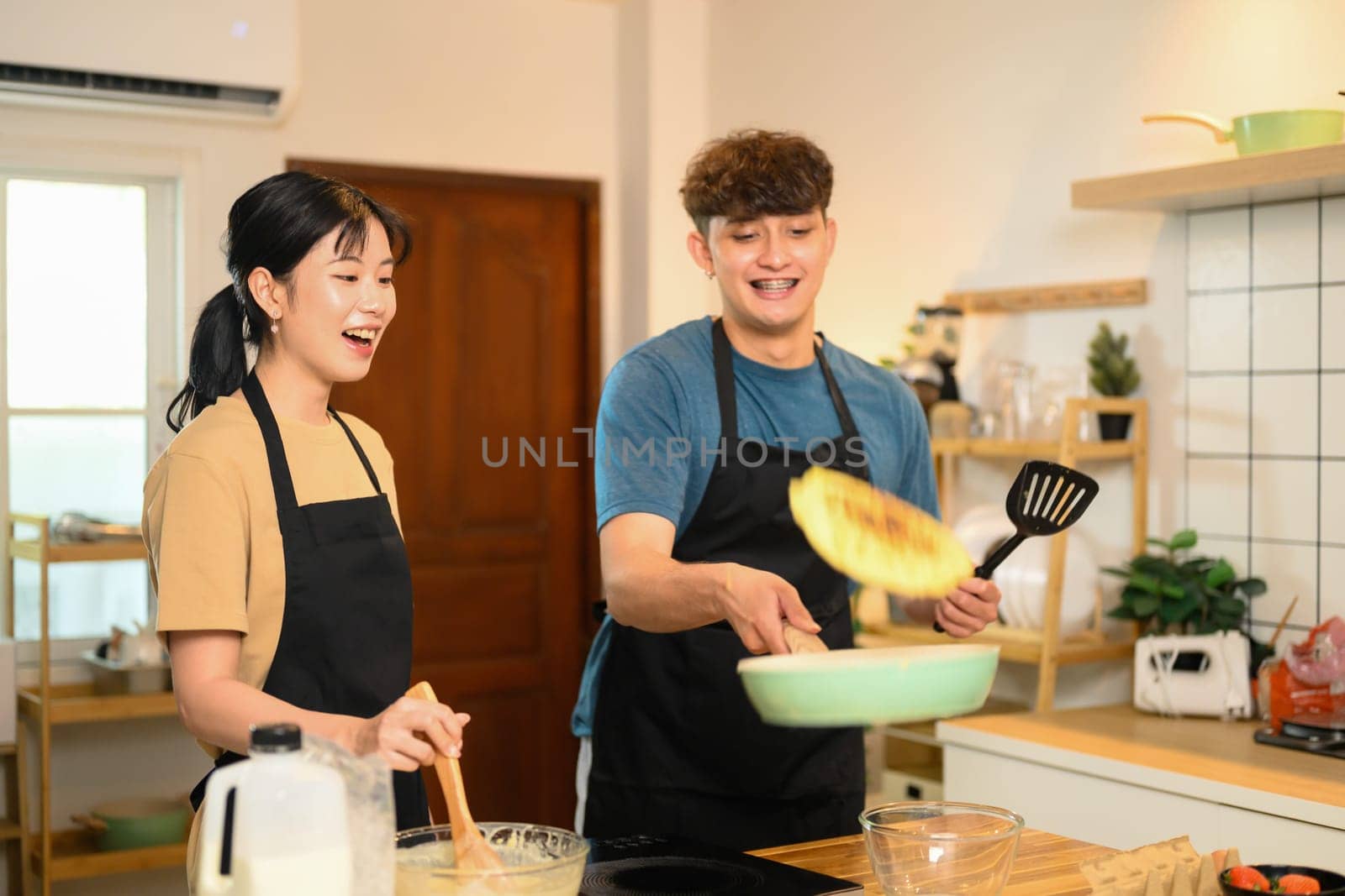 Man flipping pancake in a skillet while his partner watches and laughs.