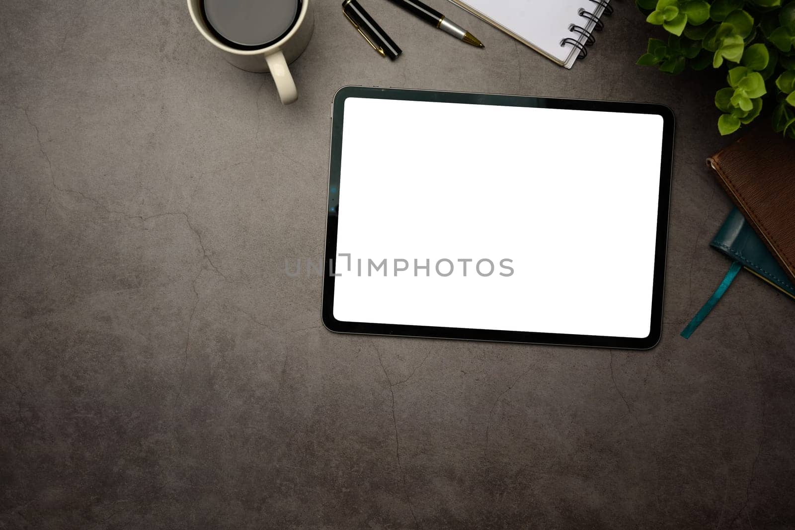 Digital tablet with blank screen on dark textured desk, surrounded by coffee cup and notebook.