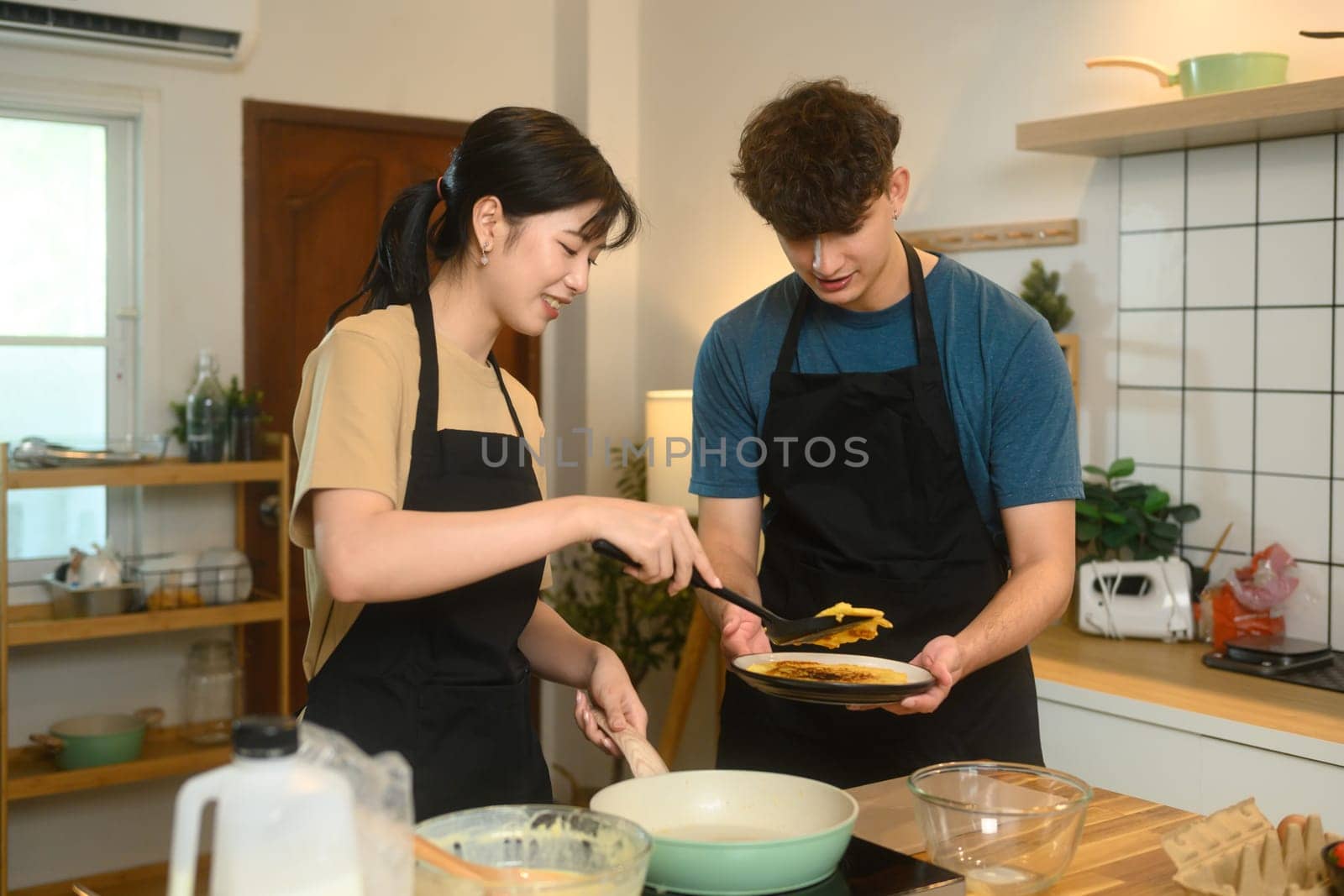 Cheerful young couple preparing breakfast pancakes in cozy home kitchen.