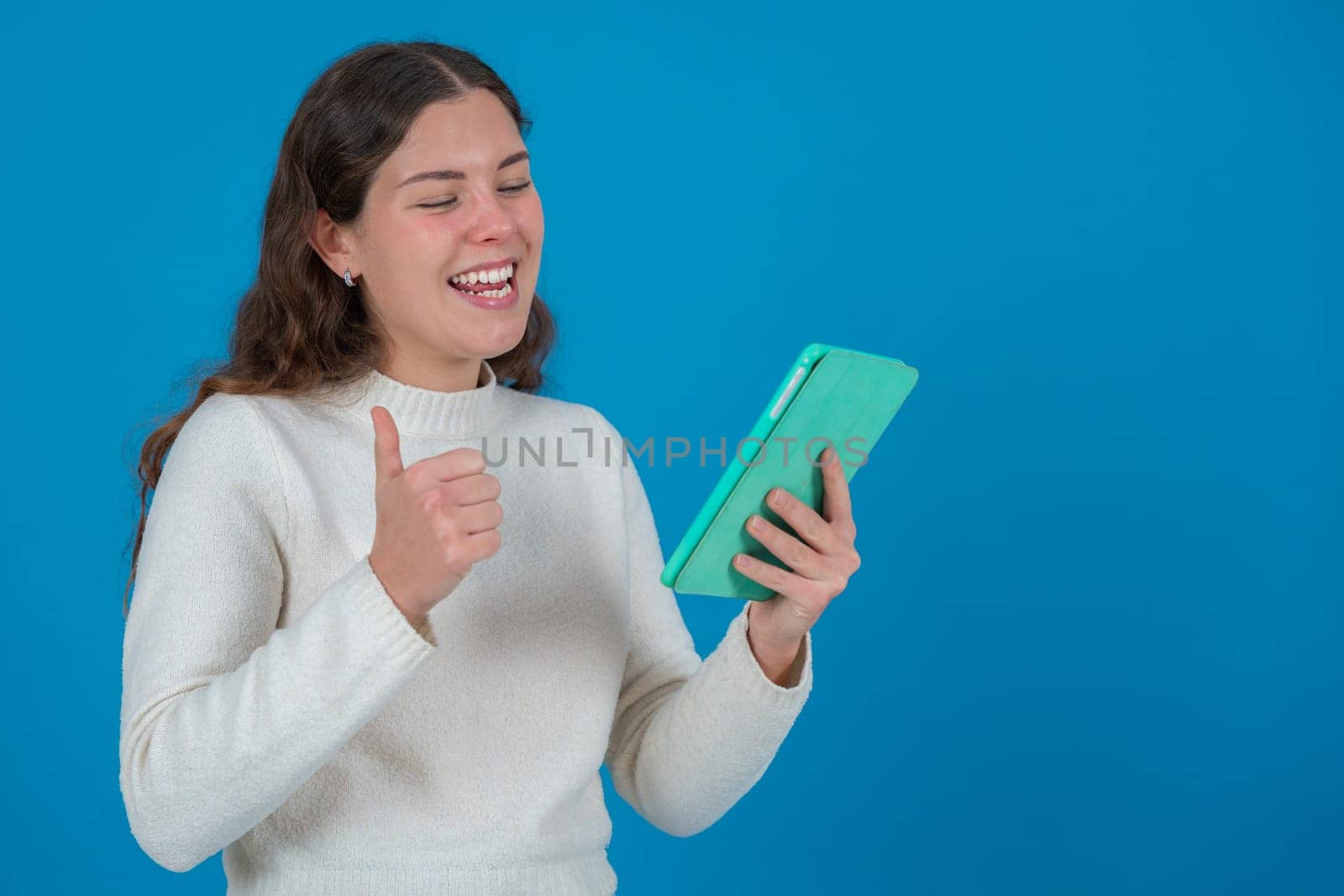 Happy young Caucasian woman holding tablet and showing thumbs up with a cheerful expression. Studio shot on blue background with copy space, ideal for communication or tech concepts.