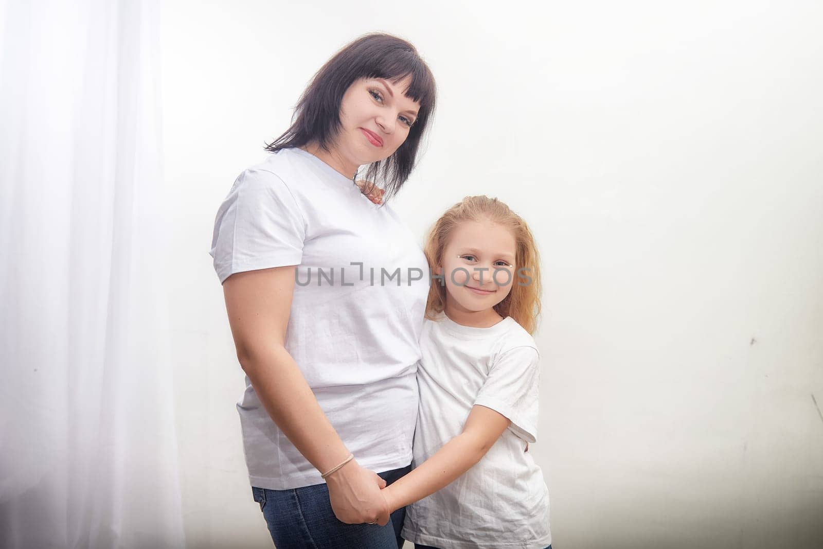 A mother and daughter stand close, holding hands and smiling. They wear matching white shirts against a bright backdrop.