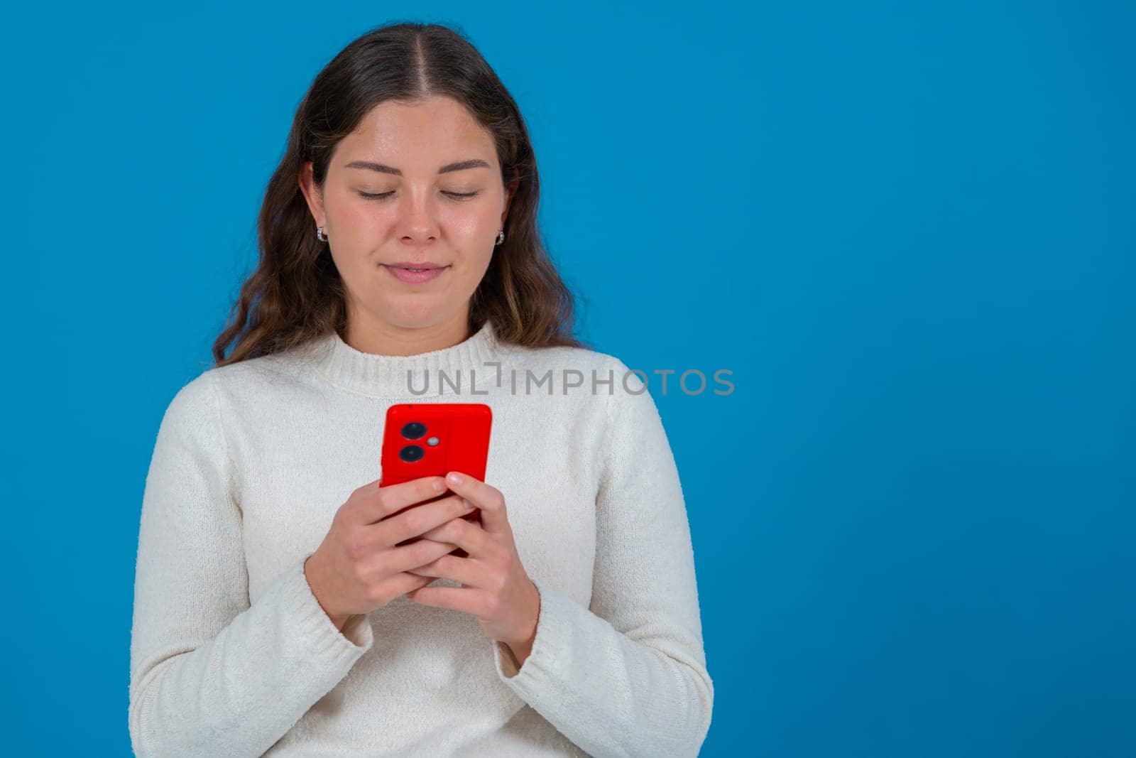 Portrait of a young woman in white sweater looking down at a red smartphone while standing against a vibrant blue background, representing focus,communication,and casual digital lifestyle..Copy space
