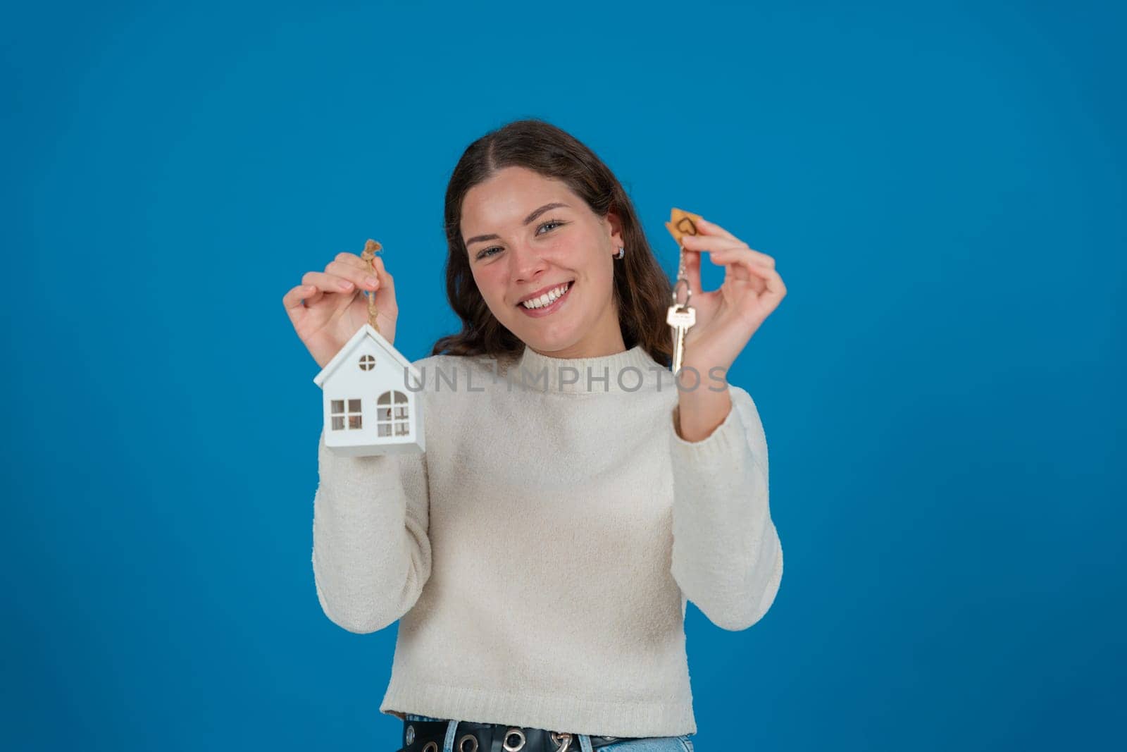 Happy young woman holding a house model and keys, expressing excitement. She wears a white sweater and stands against a blue background. Concept of home ownership, real estate, or investment. Copy space