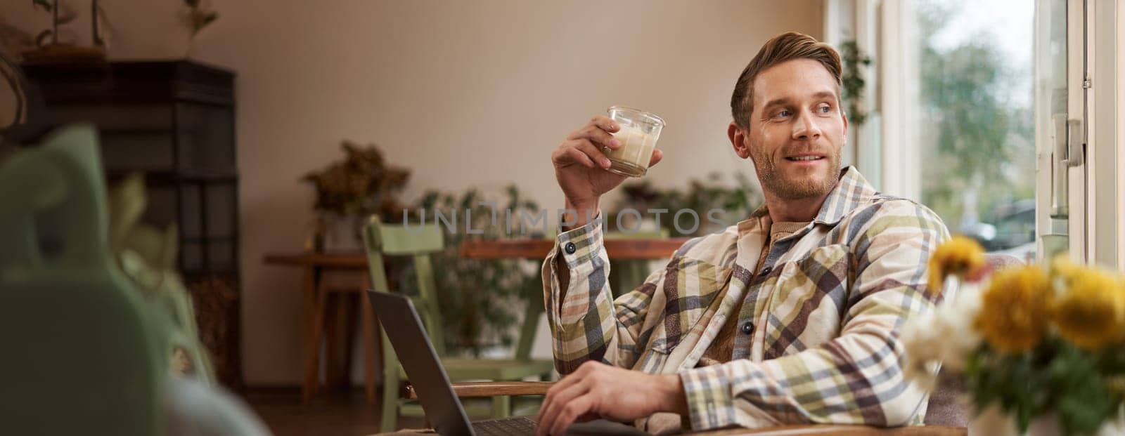 Portrait of young freelancer, a man in casual clothes, sitting in cafe and drinking coffee, working remotely on laptop, digital nomad doing his project outdoors in co-working space.