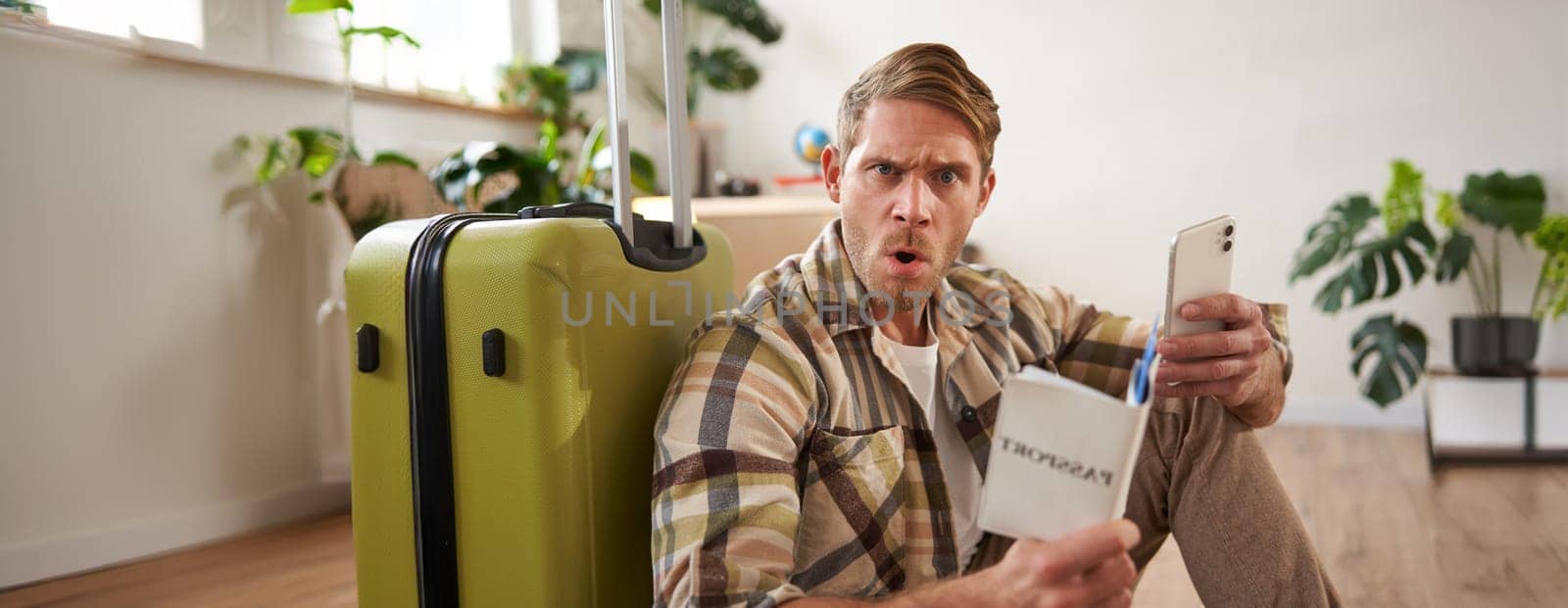 Man looks very confused, using smartphone, holding passport, sitting near luggage, planning his journey or holiday. Copy space