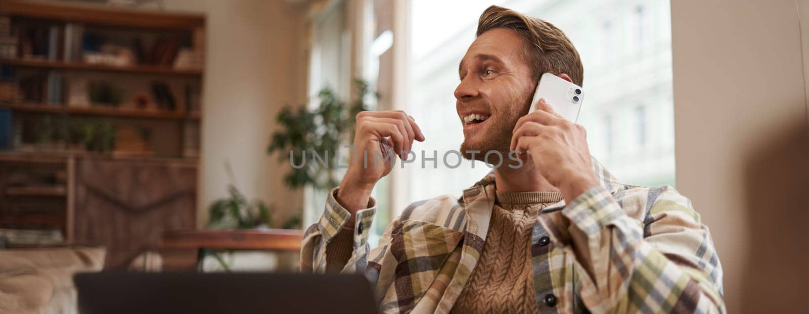 Portrait of young businessman, freelancer working remotely from coffee shop in city, sitting in chair with laptop, answer a phone call, talking on mobile to someone.