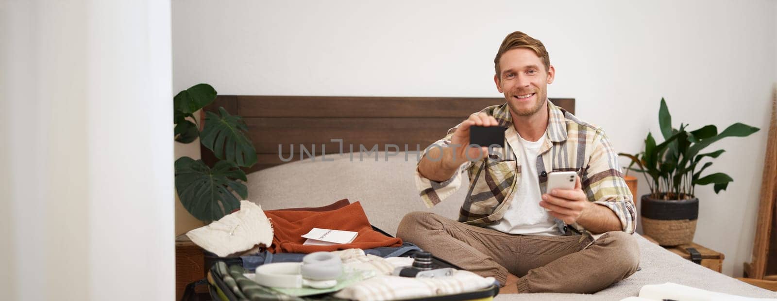 Portrait of happy man buying tickets with credit card cashback, sitting on bed and packing clothes for holiday, putting items in suitcase, holding mobile phone.