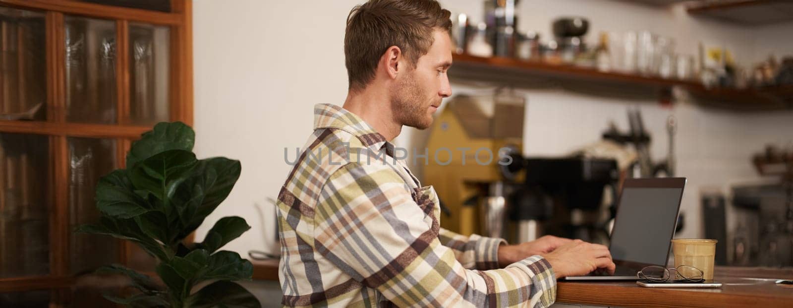 Portrait of young man, digital nomad working on project, using laptop in cafe, drinking coffee and typing, browsing on the internet.