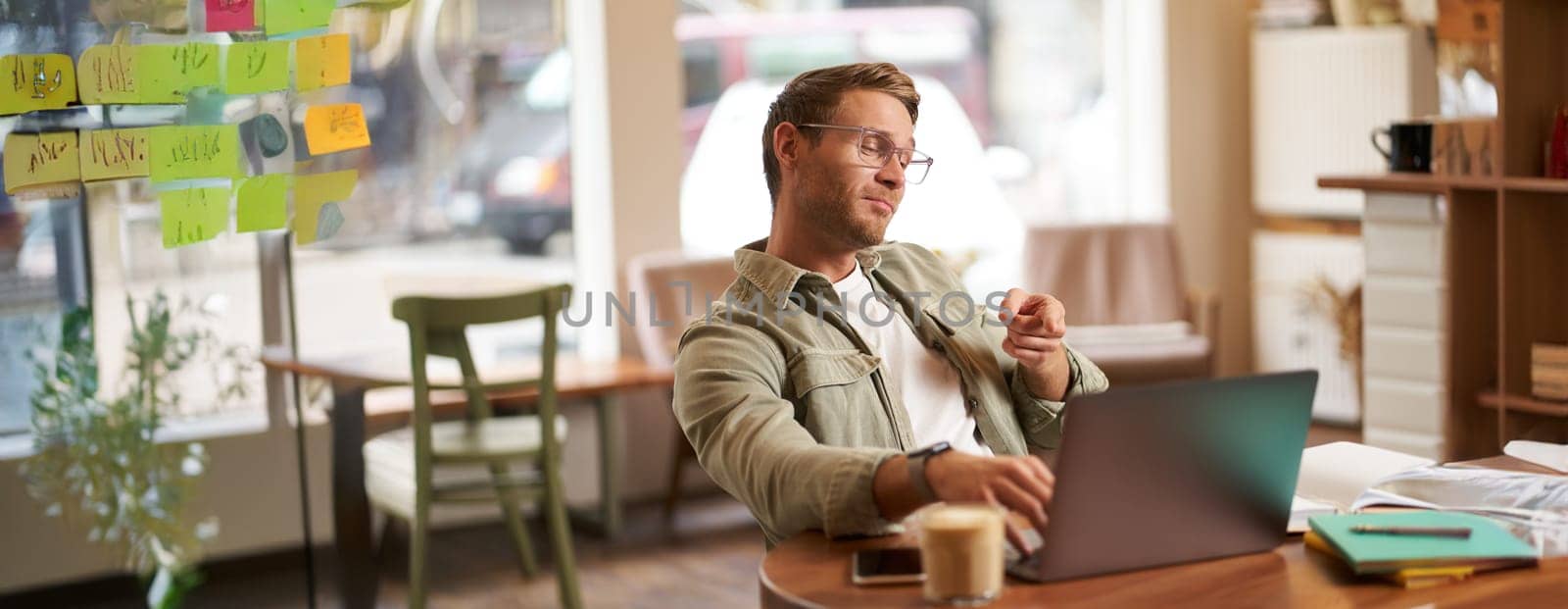 Portrait of handsome man in glasses, sits in cafe, points finger at laptop screen, video chats, connects to online meeting, feeling confident, working remotely.