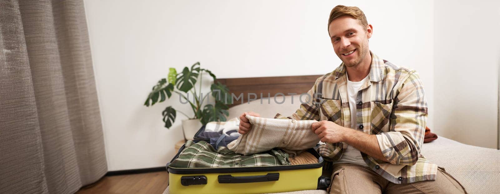 Portrait of young man sits on bed with suitcase, goes on holiday, packs clothes for vacation or business trip, checks his luggage.