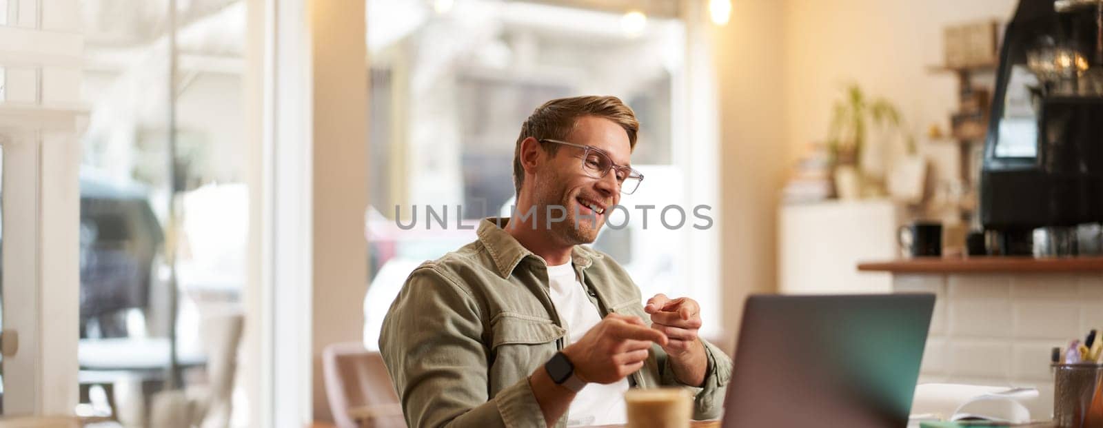 Portrait of handsome, smiling young man, online tutor, businessman working in cafe remotely, waving hand at laptop, connects to video chat or meeting.