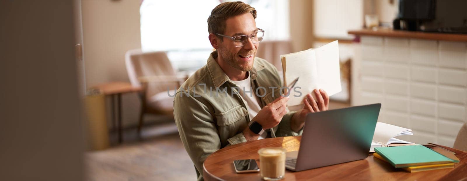 Portrait of handsome adult man showing his exercise to tutor, studying online from coffee shop, sitting in cafe with laptop, pointing at sentence in notebook.