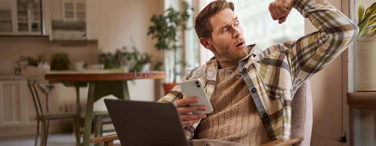 Portrait of young athletic guy sitting in cafe with laptop and smartphone, showing his biceps, flexing muscles and looking pleased at himself.