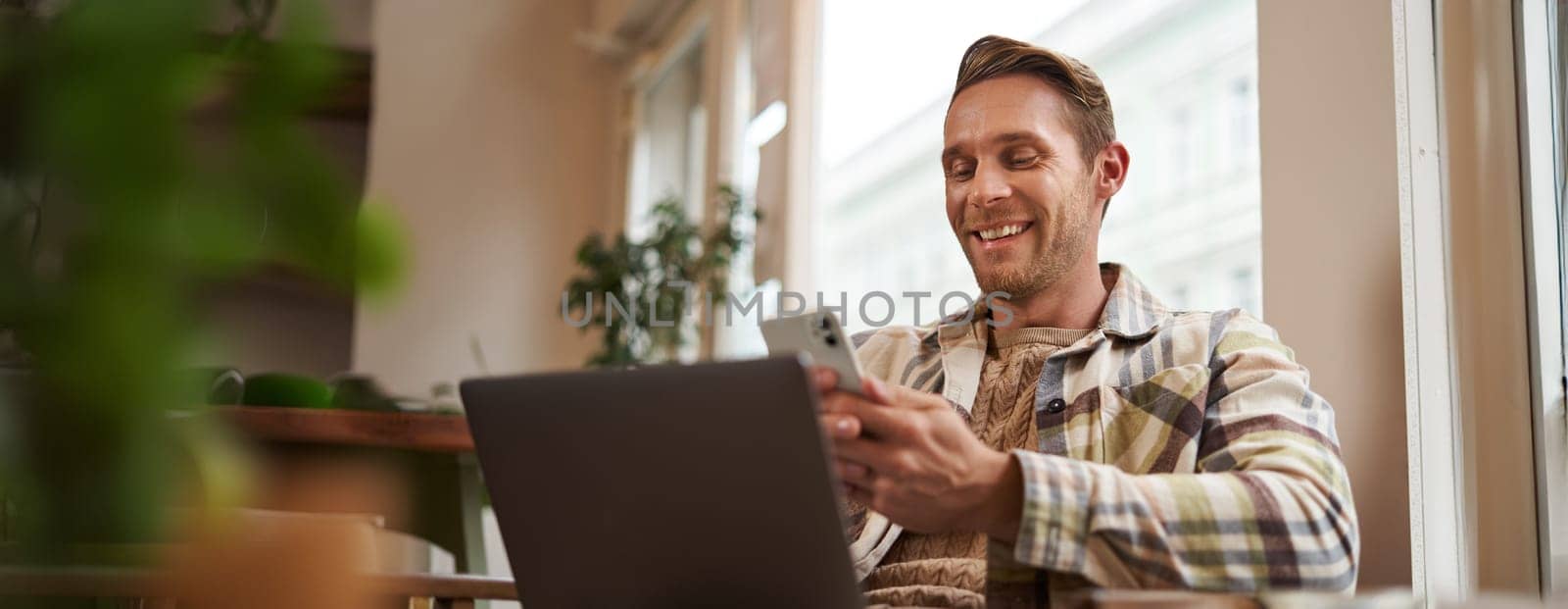 Close up portrait of handsome smiling man with laptop, cafe visitor sitting in chair, using smartphone, looking happy, messaging with friends while enjoying cup of coffee.