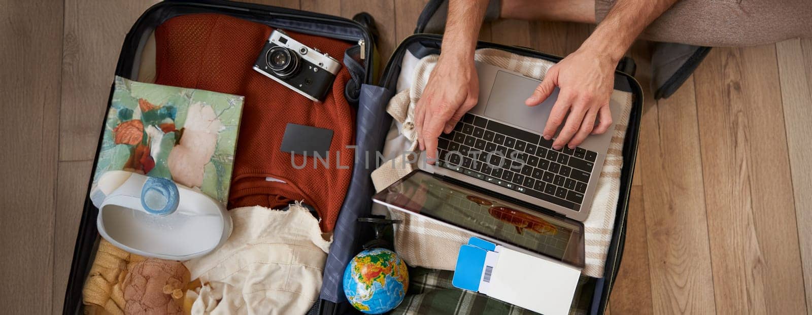 Close up portrait of male hands using laptop on top of opened suitcase, man packing for holiday, tourist with globe, booking vacation on travel agency website, picking a tour. Tourism concept