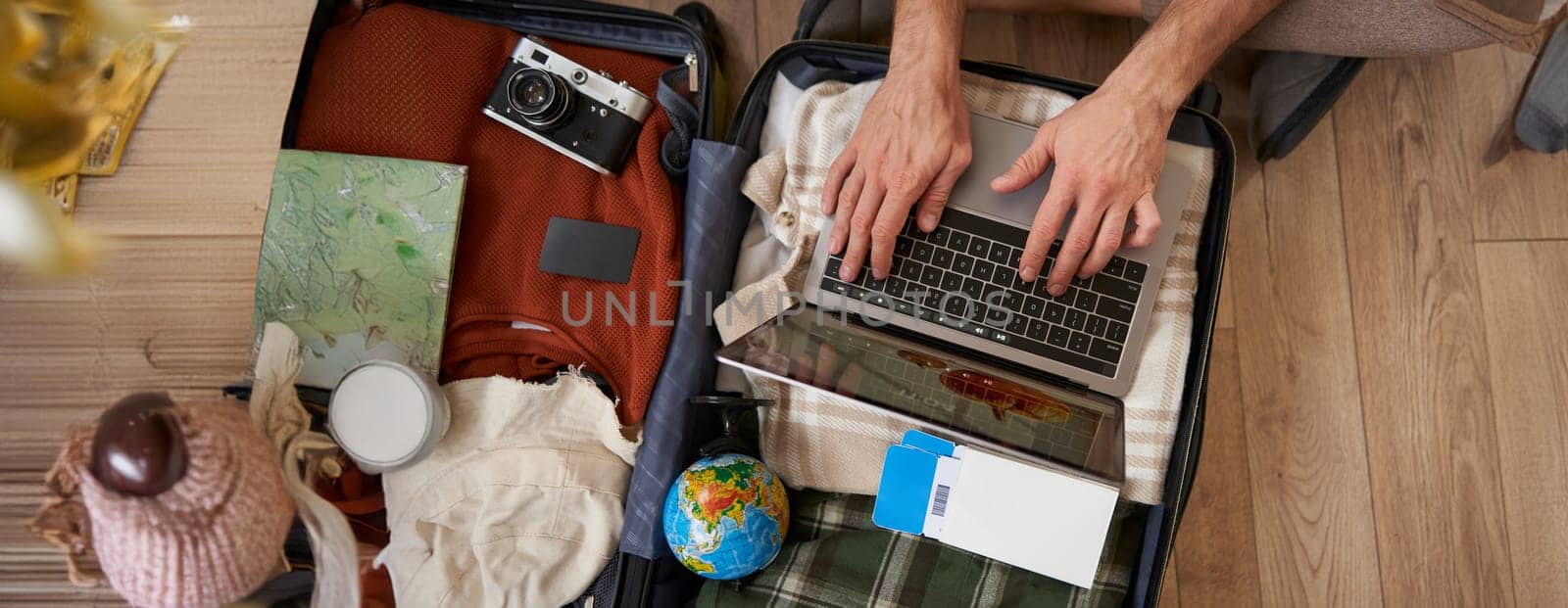 Close up of tourist, hands typing on laptop, opened suitcase with clothes for holiday, man packs luggage.