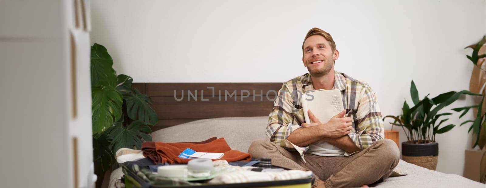 Portrait of thoughtful young man, tourist planning his vacation, sitting with open suitcase on bed, making notes, writing to do check list in notebook.