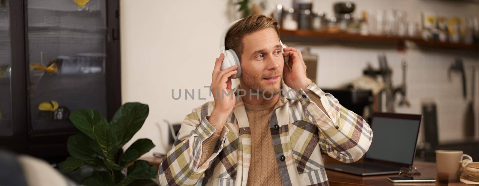 Lifestyle portrait of happy, excited young man, sitting in coffee shop with laptop and headphones, dancing on his chair and enjoying great quality sound, streaming favourite songs.