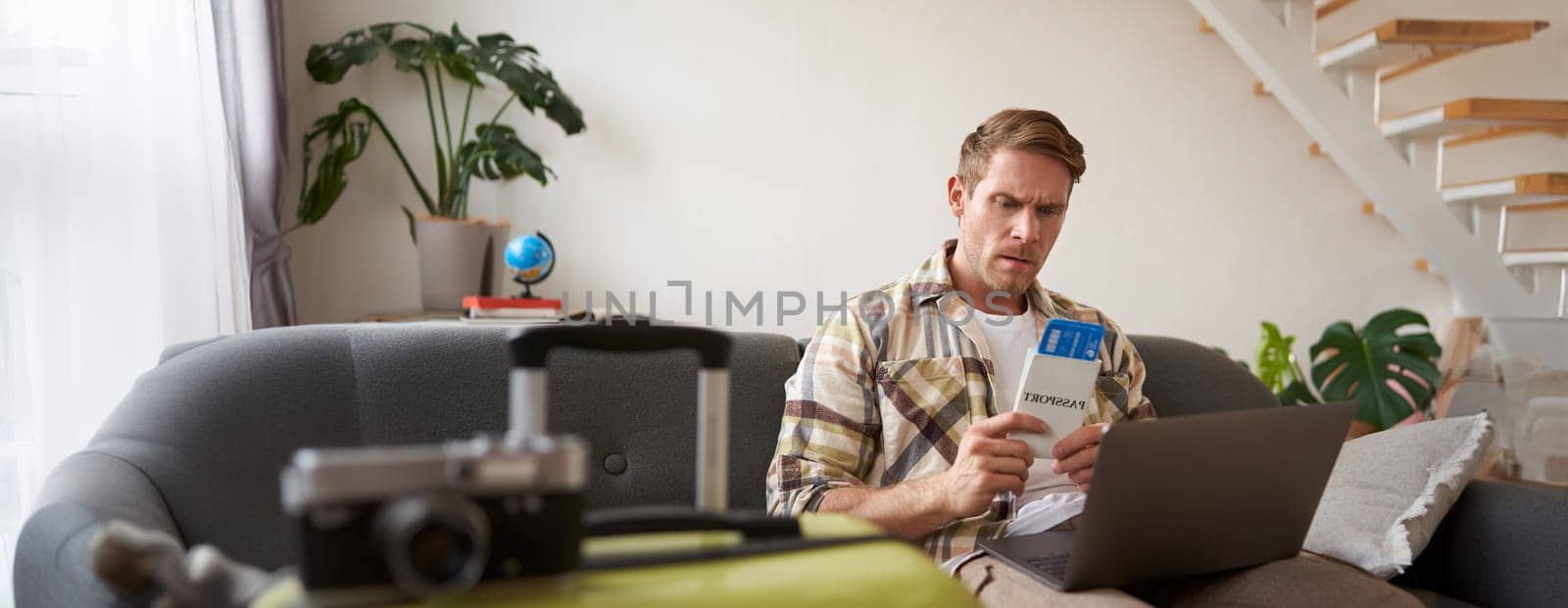 Portrait of grumpy man, holding tickets and passport, has laptop opened, looking concerned and angry, sitting with suitcase on sofa.