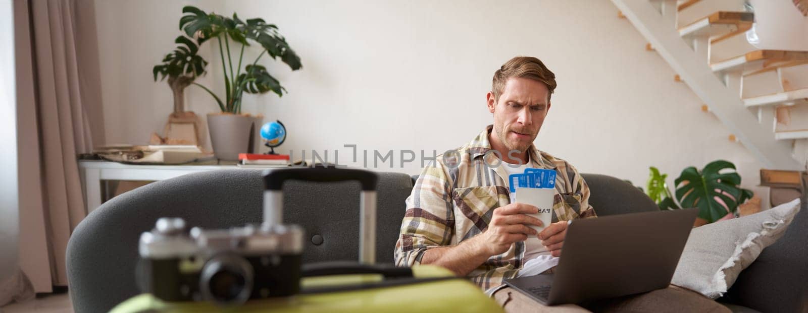 Portrait of frowning man, looking concerned at his flight tickets and passport, has laptop opened on his laps.