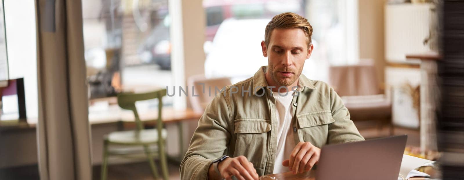 Image of young man, concentrated on his task, sitting with laptop in cafe, putting on glasses.
