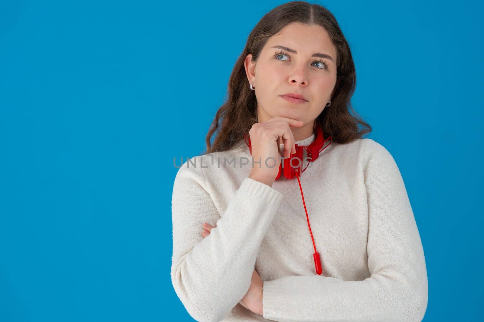 Pensive young woman with red headphones thinking and looking up, standing against a solid blue background. Concept of ideas, decision-making, and imagination with space for text or design.