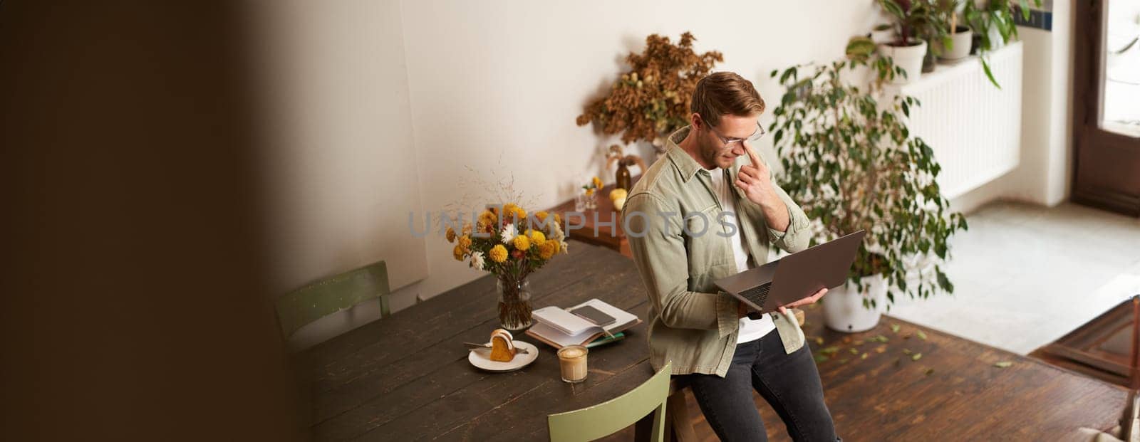 Image of young man, successful businessman sitting on table, looking at laptop, working in an office, looking concentrated on the project, doing job task, drinking coffee.