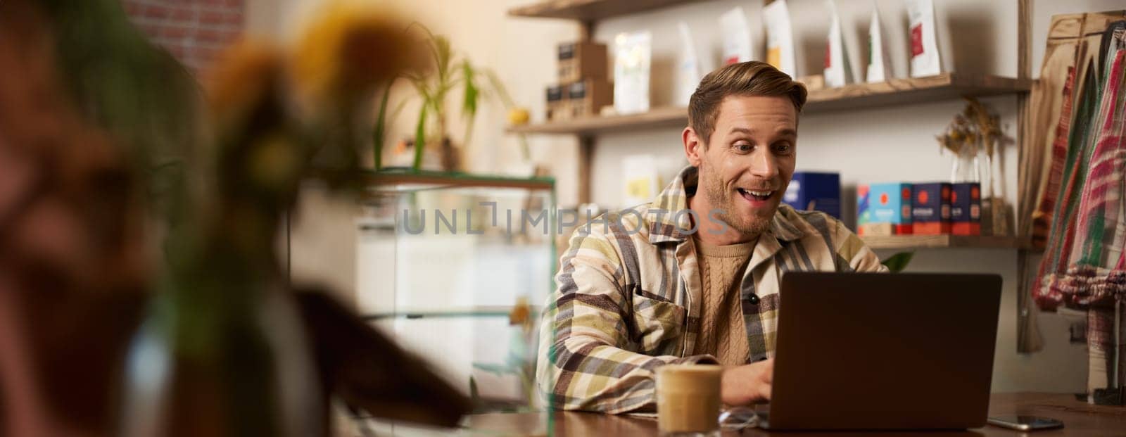 Portrait of handsome young digital nomad, man working in cafe on laptop, looking happy and pleased with his online project, sitting in co-working coffee shop.