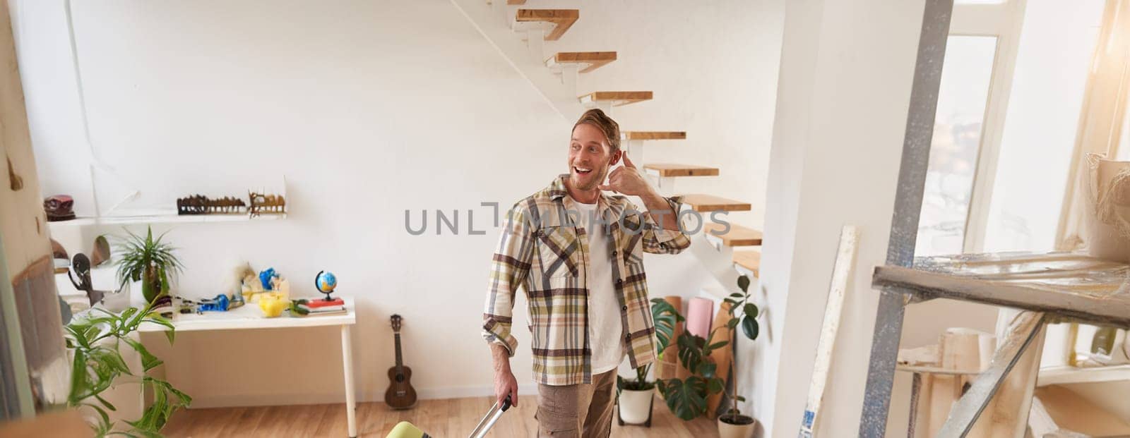 Portrait of handsome young man going on a trip, shows mobile phone hand sign gesture, going on vacation, leaving the apartment with suitcase, carrying luggage in hand.