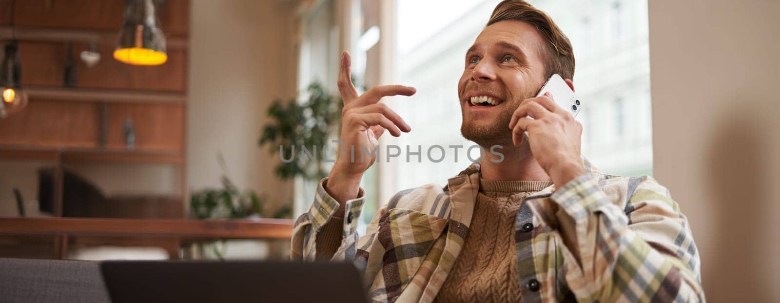 Lifestyle shot of young happy man sitting in cafe with laptop, answer phone call, talking to friend on mobile and laughing, chatting to someone with joyful face, resting in chair.