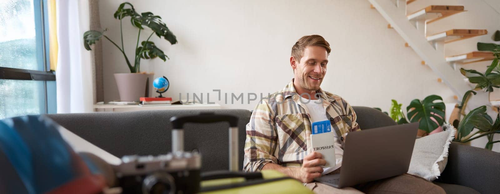 Portrait of handsome young man, has two tickets and passport, goes online on laptop, booking hotel and planning his holiday, sitting in living room.
