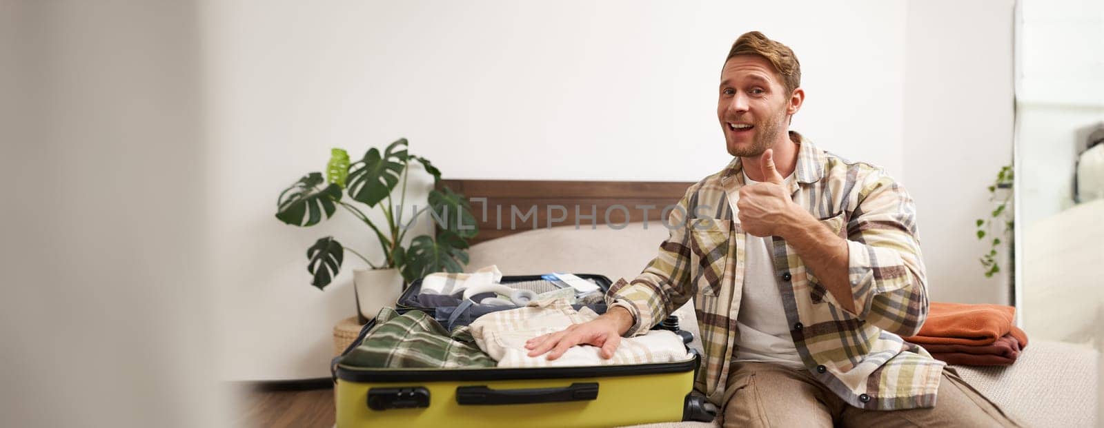 Attractive young man smiling, showing thumbs up, showing his clothes inside suitcase, getting ready for a holiday, travelling with travel agency tour. Lifestyle and tourism concept