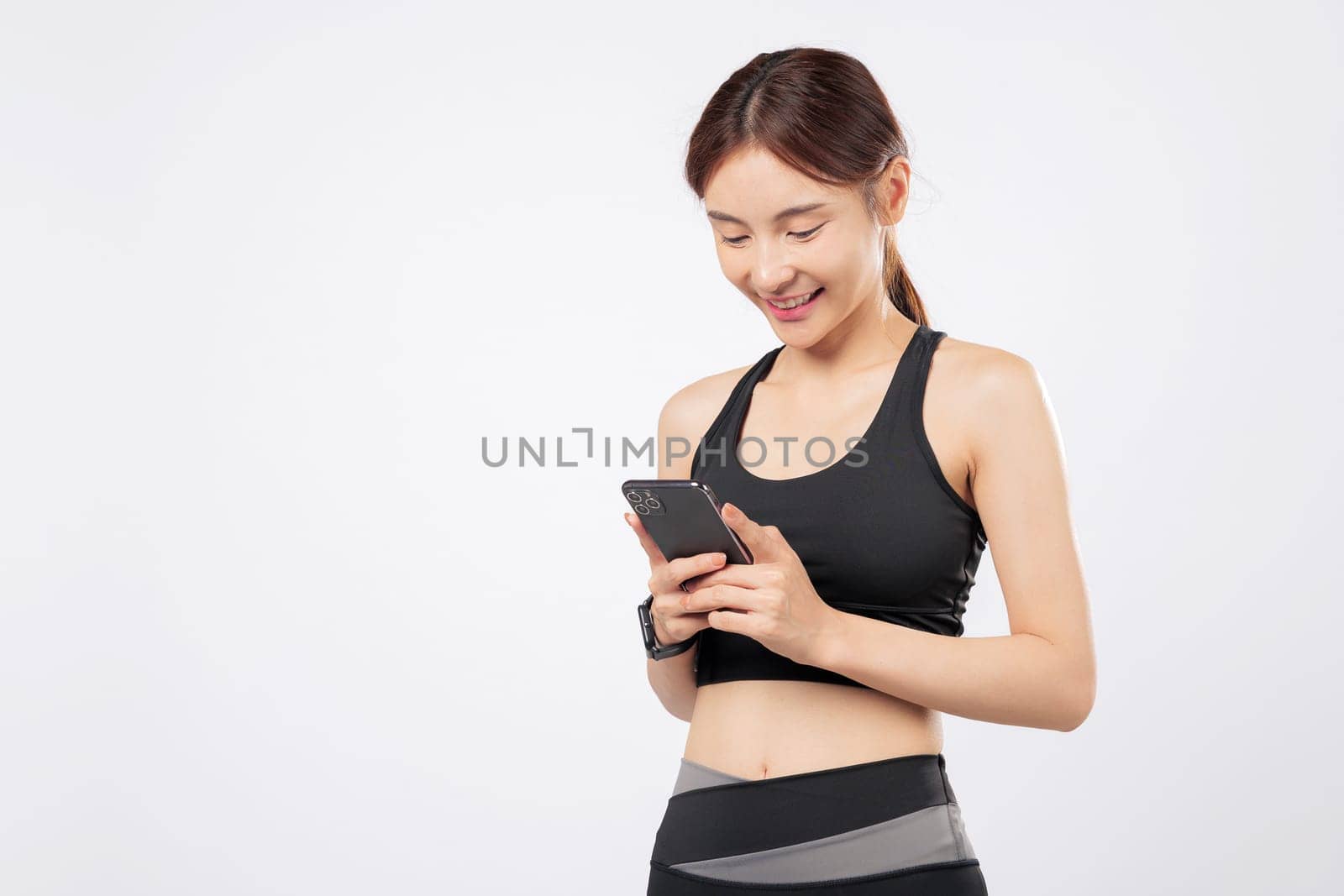With her phone ready and her phone open to a workout app, this strong woman smiles confidently in sporty attire, blending gym life and modern mobile technology. isolated studio white background
