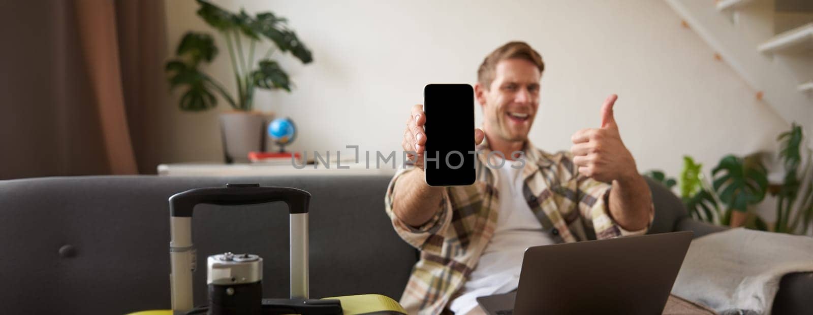 Portrait of man, tourist shows his mobile phone screen, recommends travel app, sitting with suitcase and laptop.