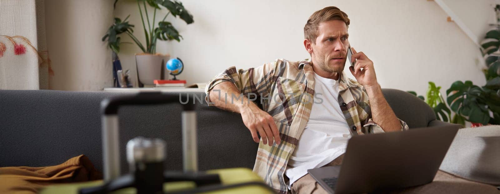 Portrait of young man with credit card, sitting and using laptop, making a phone call, booking hotel or tickets, confirming his purchase over telephone, has suitcase near him.