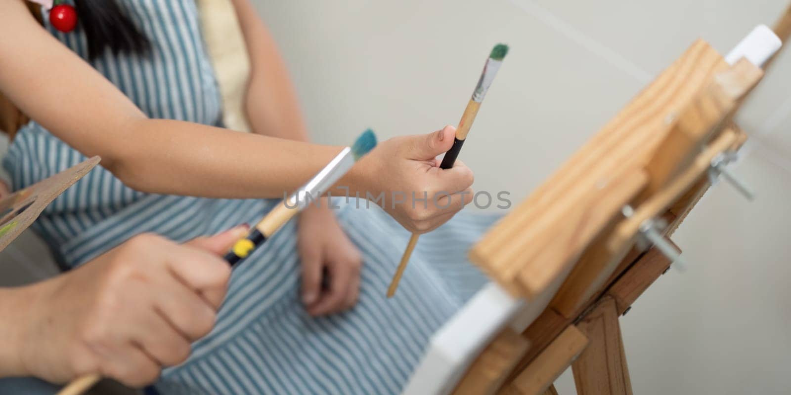 A mother supports her daughter as they hold painting brushes together, encouraging creativity and teamwork.